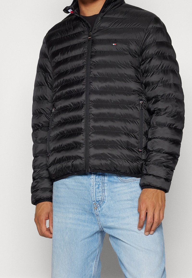 Musta toppatakki, jossa on vaakasuorat ompeleet, korkea kaulus, edessä vetoketju ja sivutaskut, päällä vaaleansiniset farkut.