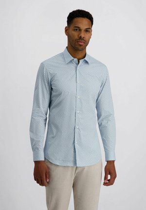 Mann steht vor einfachem Hintergrund, trägt ein hellblaues, langärmeliges Hemd mit kleinen dunklen Punkten und eine beige Hose.