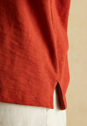 Rostfarbenshirt mit Kurzarm aus strukturiertem Stoff und einem Schlitz am Saum. Verfügt über kontrastierende Nähte und eine lockere Passform.