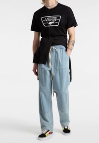 Svart grafisk t-shirt med vit logotyp, ljusblå vida denimbyxor och svarta skor med röda och gula detaljer på sidorna.