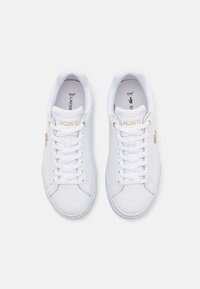 Vita sneakers i ett mjukt material, med guldaccenter, traditionella snören och "LACOSTE"-logotypen. Minimalistisk design med rundade tår.