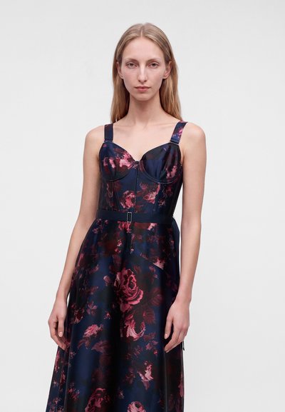Jeune femme aux longs cheveux blonds porte une robe bleu marine avec des motifs floraux roses et des bretelles épaisses, debout devant un fond uni.