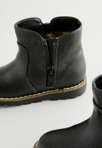 Schwarze Knöchelschuhe aus strukturiertem Kunstleder, mit einem Reißverschluss, kontrastierenden Nähten, einer robusten braunen Sohle und abgerundeten Zehen.
