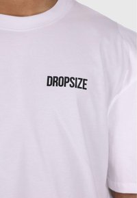 Hvit bomulls t-skjorte med en svart "DROPSIZE" logo trykket på venstre brystområde. Klassisk rund hals og korte ermer.