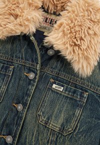 Giacca di jeans con colletto in morbido pelliccia sintetica beige. Presenta bottoni in metallo e una piccola etichetta con il marchio sulla tasca anteriore. Tessuto blu scolorito.