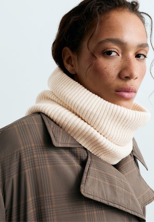 Une femme aux cheveux foncés porte une épaisse écharpe en tricot couleur crème et un manteau à carreaux bruns. Elle regarde l’objectif sur un fond clair.