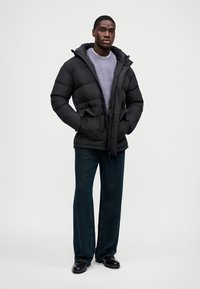 Μαύρο μπουφάν puffer με κουκούλα, δύο μπροστινές τσέπες και κλείσιμο με φερμουάρ. Φορεμένο πάνω από γκρι πουλόβερ, με σκούρο τζιν και μαύρα παπούτσια.