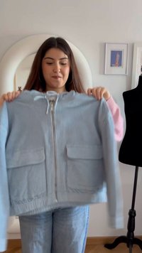 Mladá žena drží svetlomodrú bundu na zips s prednými vreckami v jasnej miestnosti so zrkadlom a čiernym manekýnom nablízku.