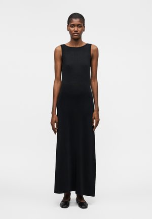 Mujer de pie, vestida con un vestido largo negro sin mangas y zapatos planos negros, frente a un fondo gris claro liso.