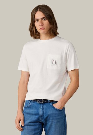 Joven con pelo castaño hasta los hombros, vistiendo una camiseta blanca con bolsillo delantero y vaqueros azules, de pie frente a un fondo liso.
