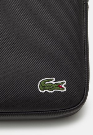 Personne portant un pull gris et un pantalon à fines rayures avec un sac bandoulière noir avec un petit logo crocodile vert et blanc.