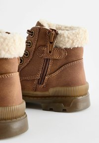 Bottes de cheville en cuir marron avec doublure en fausse fourrure beige, fermetures à lacets et à glissière, semelles en caoutchouc texturé et chevilles rembourrées pour le confort.
