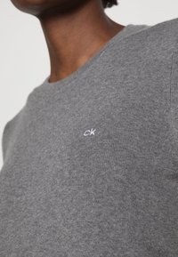 Maglione grigio a maglia con scollatura rotonda. Presenta una leggera costina e un piccolo logo "ck" bianco sul lato sinistro del petto. Texture morbida.