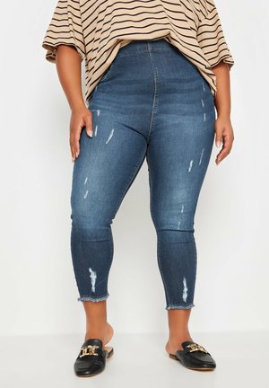 Vrouw draagt versleten blauwe skinny jeans met rafelige zomen, een beige gestreept topje en zwarte instap loafer schoenen met gouden kettingdetail.