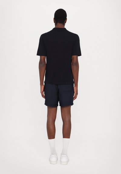 Camisa polo de manga curta azul-marinho combinada com calções azul-marinho, meias brancas e ténis brancos, vista pela parte de trás contra um fundo liso.
