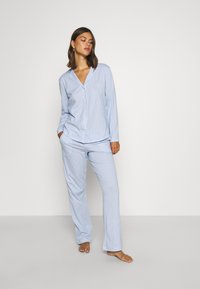 Šviesiai mėlyna dryžuota pižamų komplektas, kuriame yra mygtukais segamas ilgomis rankovėmis viršus ir atitinkamos kelnės. Pagaminta iš lengvos medžiagos, laisvas kirpimas.