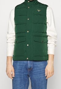 Zelená nadýchaná vesta s vysokým límcem, zapínáním na cvoky a dvěma předními kapsami. Hladký povrch a rovnoměrné švy po celé délce.