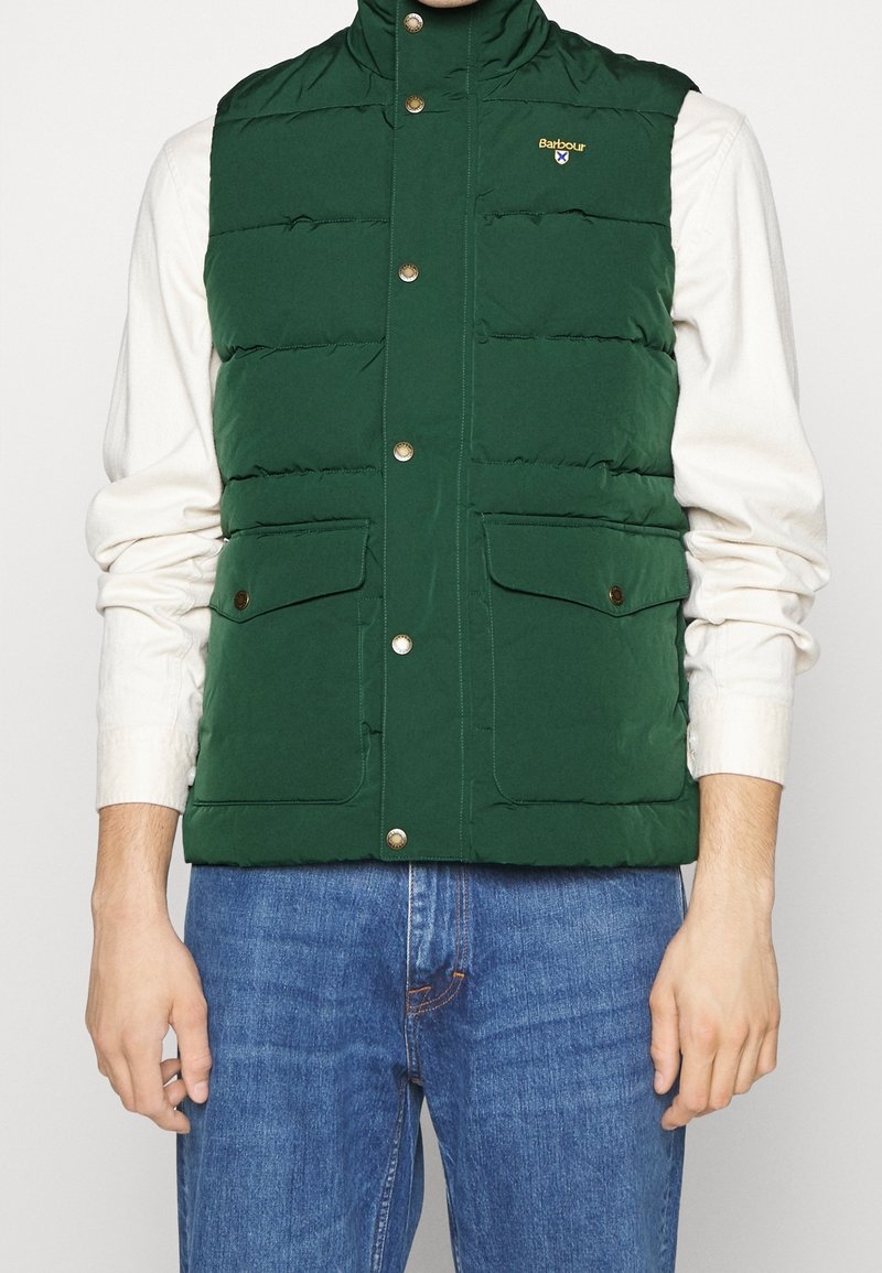 Zelená nadýchaná vesta s vysokým límcem, zapínáním na cvoky a dvěma předními kapsami. Hladký povrch a rovnoměrné švy po celé délce.