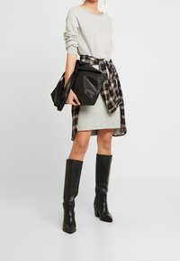 Maglione grigio a maniche lunghe sovrapposto a un vestito grigio a coste, camicia a quadri bianca e nera legata in vita, stivali neri in pelle fino al ginocchio e borsa nera.