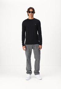 Mustade pikad varrukad shirt, hallid teksad taskutega ja valged sneakerid. Kandjal on prillid ja keskmise pikkusega pruunid juuksed.