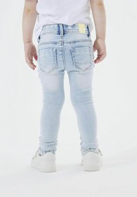 Jeans skinny de mezclilla ligera azul con un acabado desgastado, que cuentan con dos bolsillos traseros y una etiqueta amarilla en la cinturilla. Zapatillas blancas visibles.