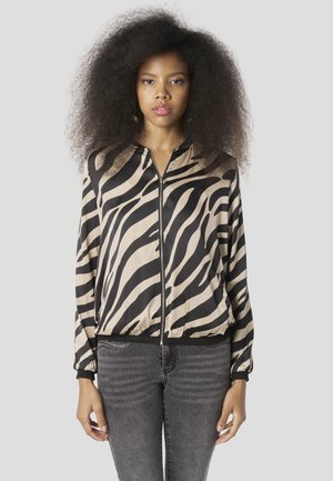 Bézs és fekete színű selyem zebra mintás bomber dzseki cipzáras lezárással, rugalmas mandzsettákkal és szegéllyel. A modellen szürke farmernadrág látható.