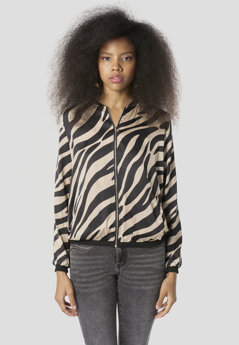 Zebra-print bomberjack in beige en zwart zijde met ritssluiting, elastische boorden en zoom. Model draagt grijze jeans.
