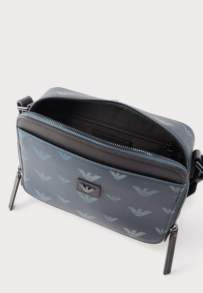 Μικρή μπλε τσάντα χιαστί με επαναλαμβανόμενο μοτίβο λογότυπου αετού, εμπρόσθια τσέπη με φερμουάρ και ανοιχτό κύριο διαμέρισμα που αποκαλύπτει την εσωτερική τσέπη με φερμουάρ.