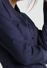 Granatowa koszula z długim rękawem o gładkiej strukturze i subtelnym połysku. Posiada kołnierzyk i czarne guziki.