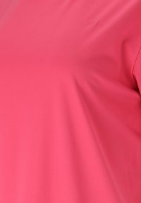 Nahaufnahme von glattem, leuchtend pinkem Stoff mit dezentem, geprägtem Logo oben rechts, das Textur und leichte Falten zeigt.
