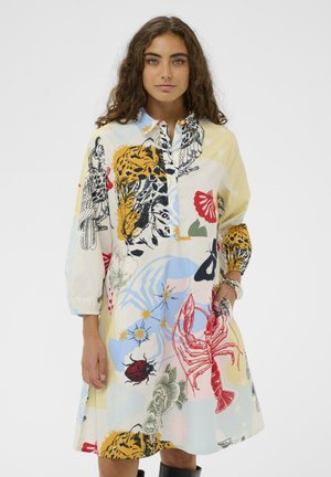 Kobieta o długich kręconych włosach, ubrana w kolorową sukienkę z nadrukami zwierząt i natury, w tym tygrysa, homara i ptaków, stojąca na białym tle.