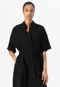 Black shirt dress made of a lightweight fabric, featuring a collar, short sleeves, a belt at the waist, and button closures along the front.
