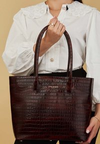Sac fourre-tout en simili-crocodile de couleur bordeaux foncé avec deux poignées, présentant une finition brillante et un accent en or avec le logo à l'avant.