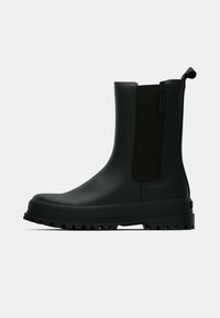 Bota preta até ao tornozelo feita de borracha lisa, com painéis laterais elásticos, sola robusta e texturizada, e uma alça na parte de trás para facilitar o calçar.