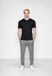 Sort t-shirt med korte ærmer, grå sweatpants med manchetter ved anklerne og hvide sneakers. Model står med hænderne i lommerne foran en ensfarvet baggrund.