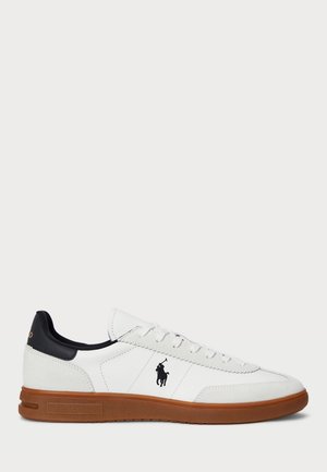 Witte lage sneaker met een bruine zool, zwarte hielaccent, veters en een zwart polo-spelerlogo aan de zijkant.
