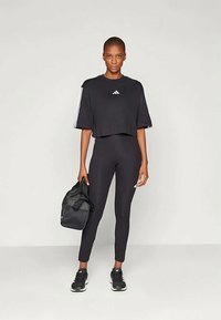 Juodas trumpas sportinis marškinėliai su balta Adidas logotipu, derinami su juodomis legginsais. Apranga pasižymi sportiniu dizainu, nešiojant juodus sportinius batelius ir laikant tamsų krepšį.