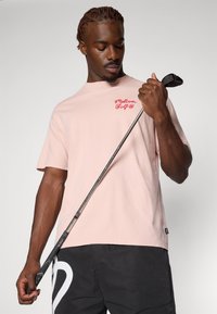 Lyserød T-shirt med rødt broderet logo, parret med sorte shorts. Teksturen ser blød ud; T-shirten har en afslappet pasform.