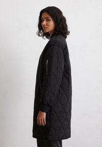 Femme aux cheveux bouclés portant un long manteau noir matelassé avec une poche zippée sur la manche, se tenant devant un mur blanc uni.