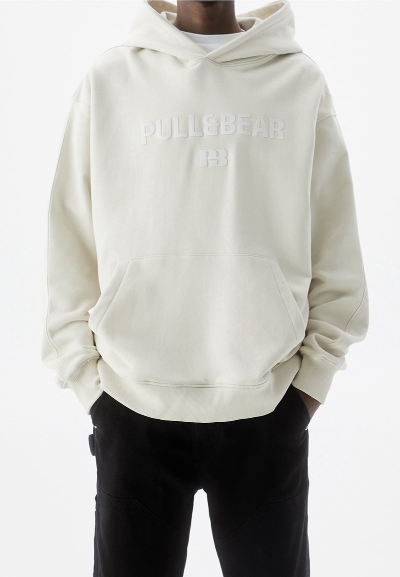 Uomo che indossa un hoodie off-white di Pull&Bear con tasca frontale e pantaloni neri, mani nelle tasche, in piedi di fronte a uno sfondo bianco uniforme.
