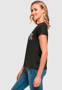 Schwarzes Baumwoll-T-Shirt mit kurzen Ärmeln, das ein grafisches Design auf der Vorderseite aufweist. Kombiniert mit blauen Jeans, die eine figurbetonte Silhouette zeigen.