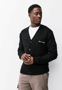 Zwarte cardigan met een V-hals, voorzien van vijf knopen en geribbelde manchetten. Geborduurd detail op de borst, zachte gebreide textuur.