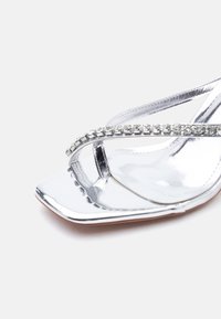 Ασημί σανδάλι με ψηλό τακούνι, τετράγωνη μύτη και λεπτό λουράκι διακοσμημένο με διαφανείς στρας σε λευκό φόντο.