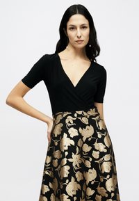 Μαύρο εφαρμοστό τοπ με κοντά μανίκια, σε συνδυασμό με μαύρη φούστα με χρυσό λουλουδένιο σχέδιο, που αναδεικνύει τη λεπτή μέση και τη ροή της σιλουέτας.