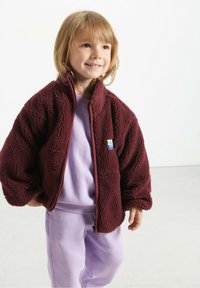 Bordo flis jakna s patentnim zatvaračem, oversized dizajna i malim zakrpama. Nosena preko lavande majice, u kombinaciji s odgovarajućim lavande hlačama.