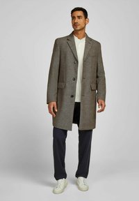 Bruine tweed lange jas met geklede revers en twee voorzakken, met zwarte knopen. Draag het over een lichtkleurig shirt en donkere broek.