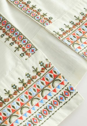 Embroidered fabric featuring a cream background, intricate patterns in red, blue, and green, with textured details on the edges.