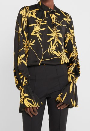 Blouse noire avec imprimé floral jaune vif, manches longues évasées et col haut noué, associée à un pantalon noir ajusté.