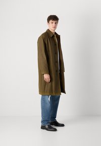 Olive green coat with a button front, long sleeves, and a straight cut. Worn over a white shirt, paired with blue jeans and black shoes.