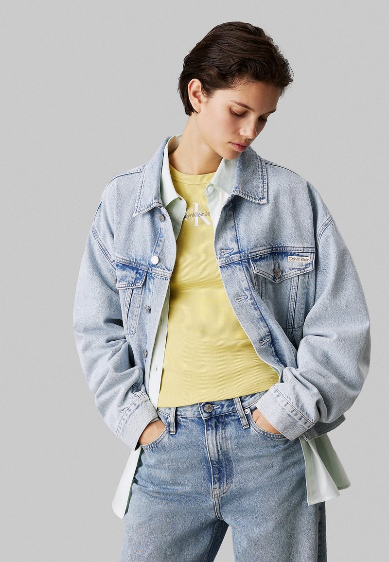 lichtblauwe spijkerjas met een relaxte pasvorm, gedragen over een gele geribbelde top. Opvallende details zijn een mintgroen shirt eronder en subtiele branding.
